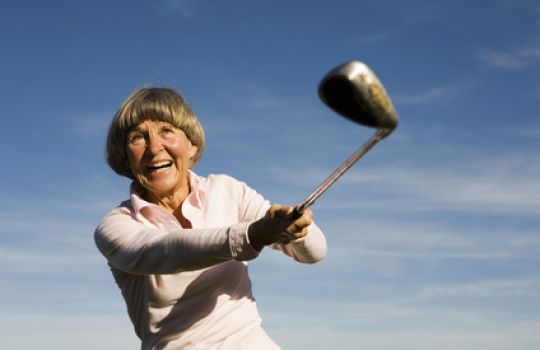 Ältere Frau hält Golfschläger in der Hand, steht vor blauem Himmel.